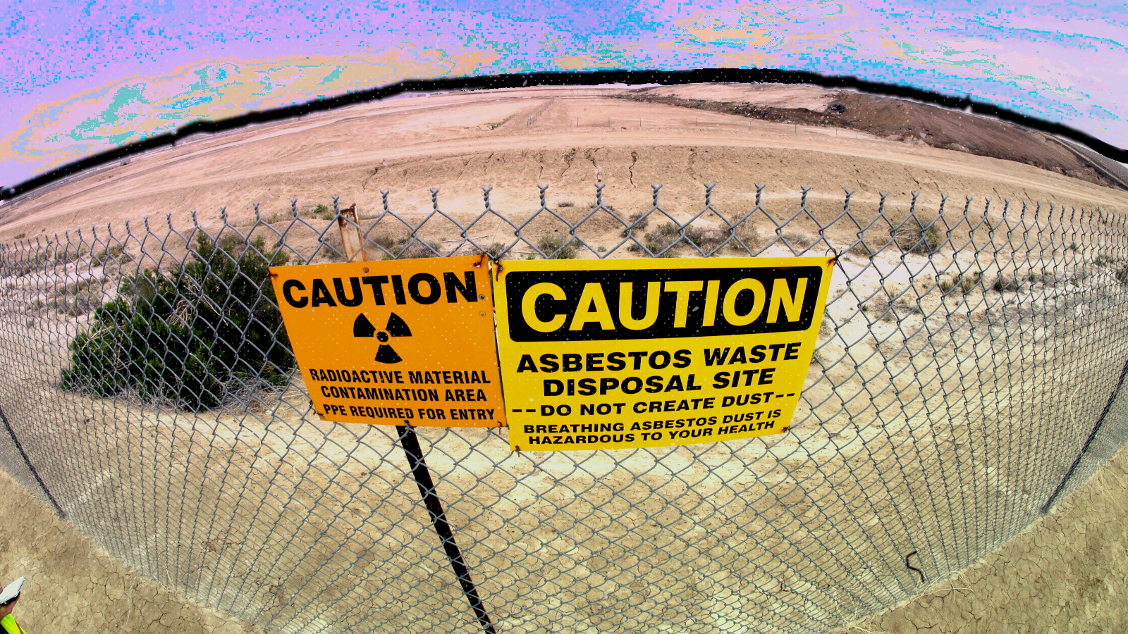 A caution sign hanging on a wire fence reads “asbestos waste disposal site.”
