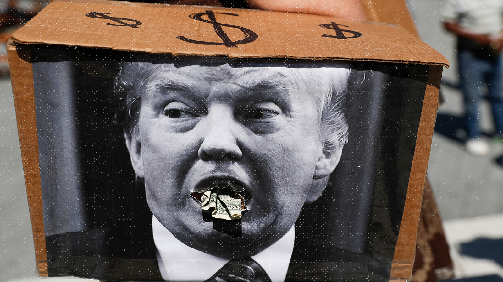 A satirical image of Donald Trump with money coming out of his mouth is pasted on top of a box covered in dollar signs.