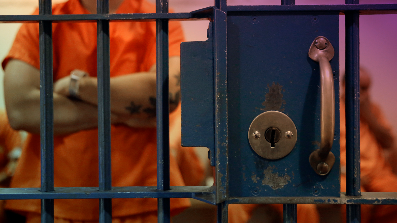 Prisoners in orange jumpsuits stand and sit in a locked holding cell.