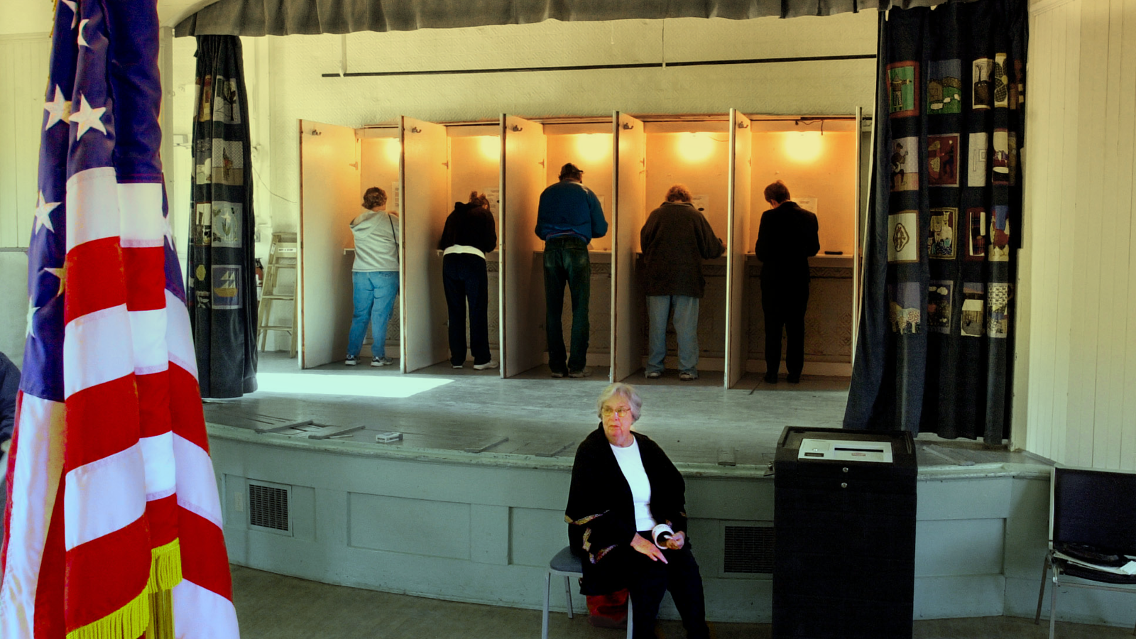 A photo of people in a voting booth in the background, with an American flag and poll worker in the foreground.